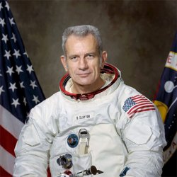 A photograph of Deke Slayton in his space suit, without the helmet, in front of 2 American flags.